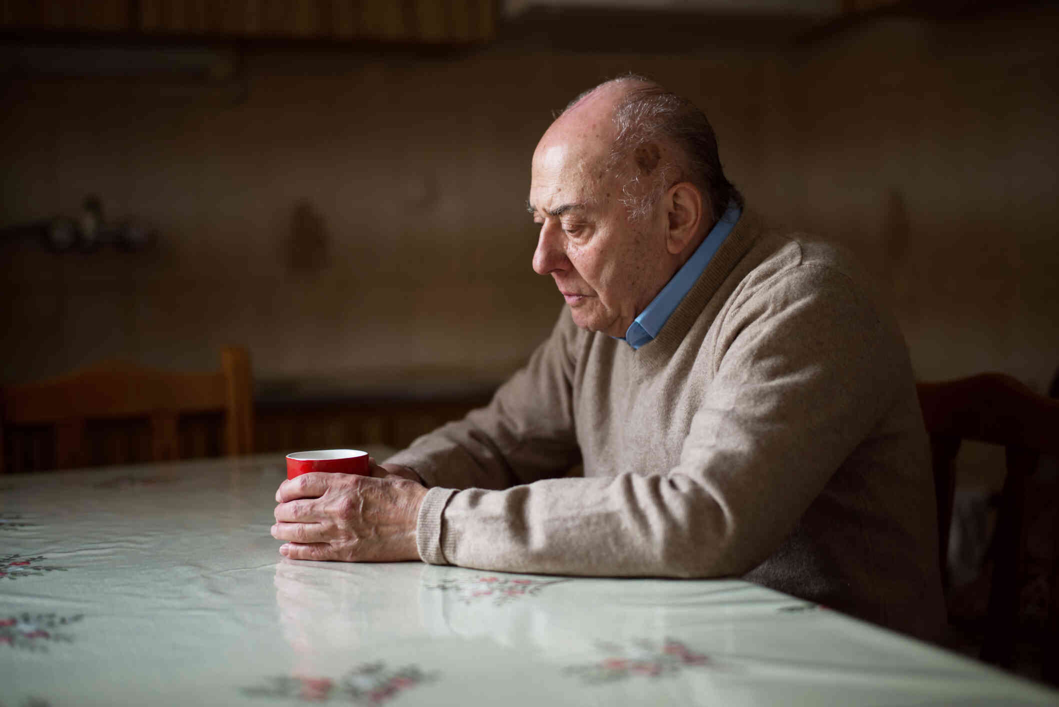 An elderly man in a tan sweater sits at his kitchen table and cups his hands around a coffee mug while gazing down sadly.